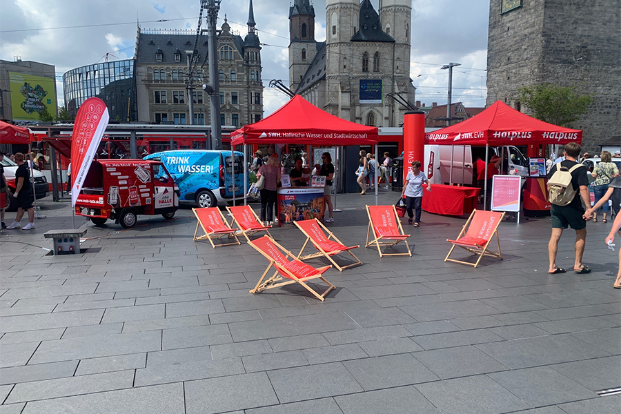 Das Bild zeigt einen Informationsstand der Stadtwerke Halle auf dem Marktplatz in Halle (Saale) mit Liegestühlen und einem Werbefahrzeug, das zum Wassertrinken aufruft. 
