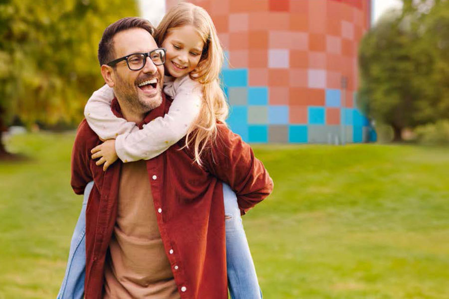 Ein Mann mit Brille trägt ein lachendes Mädchen auf dem Rücken vor einem bunt gemusterten Turm in einer Parklandschaft.