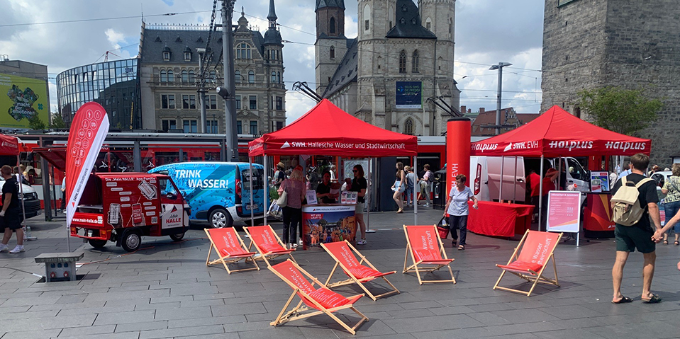 Das Bild zeigt einen Informationsstand der Stadtwerke Halle auf dem Marktplatz in Halle (Saale) mit Liegestühlen und einem Werbefahrzeug, das zum Wassertrinken aufruft. 