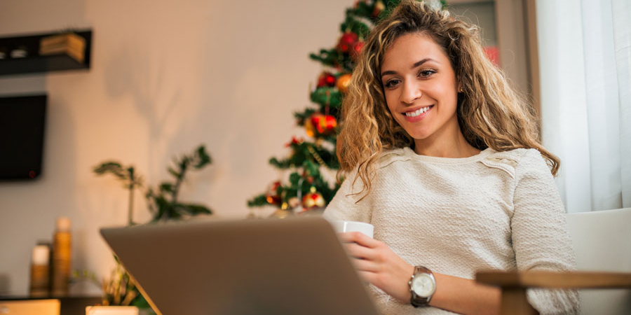 Eine junge Frau mit blondem lockigen Haar blick auf Ihren Laptop, der auf ihrem Schoß steht. In der linken Hand hält Sie eine Tasse, sie lächelt. Im Hintergrund ist ihr Zimmer zusehen, in dem auch ein Weihnachtsbaum steht.