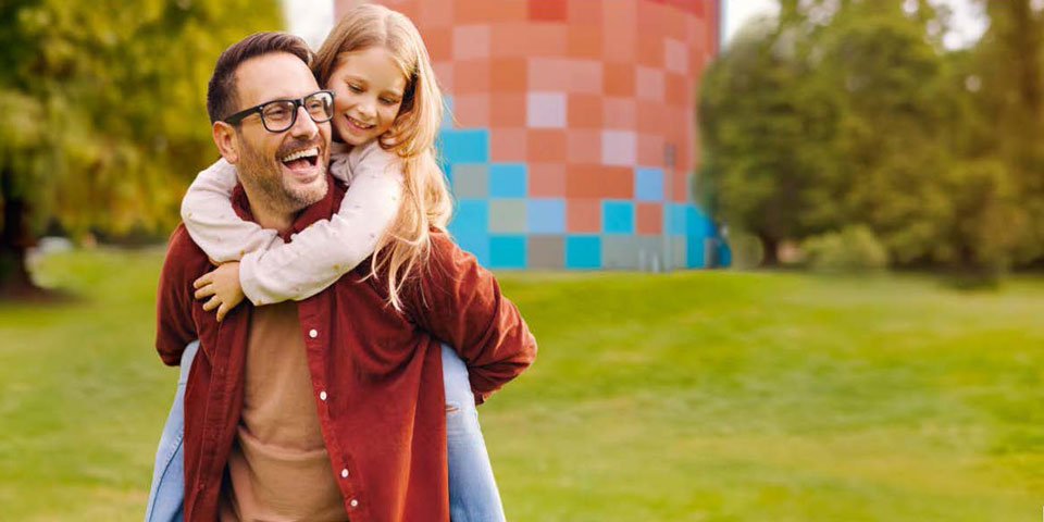 Mann mit Brille trägt ein lachendes Mädchen auf dem Rücken vor einem bunt gemusterten Turm in einer Parklandschaft.