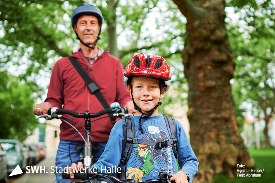 Ein Mann mit Helm und rotem T-Shirt schaut mit seinem Fahrrad in der hand Richtung Kamera. Vor ihm steht ein kleiner Junge. Er trägt ein hellblauen Pullover und einen roten Helm. Auch er hält sein Fahrrad fest. 