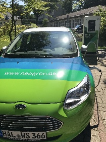 Das Bild im Hochformat zeigt ein Elektroauto. Es ist im neogrün-Design und hat die Farben grün und blau. Im Hintergrund sieht man die neue Elektroladesäule.