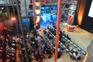 Das Bild zeigt eine Menschenmasse von oben: Viele Personen sitzen in einer großen Halle des Kraftwerk und schauen nach vorn. Dort steht in stimmungsvollem Licht Klaus Rehda und spricht zu den Personen.