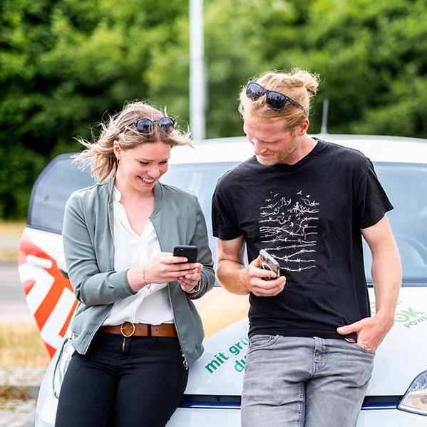 Mann und Frau lehnen am Auto und schauen auf ein Handy