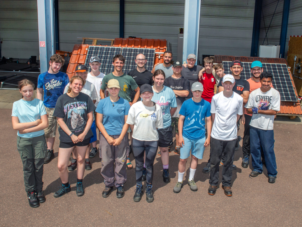 Eine Gruppe von Jugendlichen und Erwachsenen steht vor einer Werkhalle für ein gemeinsames Gruppenfoto. Im Hintergrund sind Solarmodule und Dachziegel zu sehen.