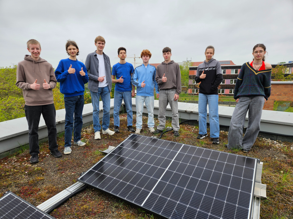 Acht Jugendliche stehen auf einem begrünten Flachdach hinter Solarmodulen und zeigen teilweise den Daumen nach oben.