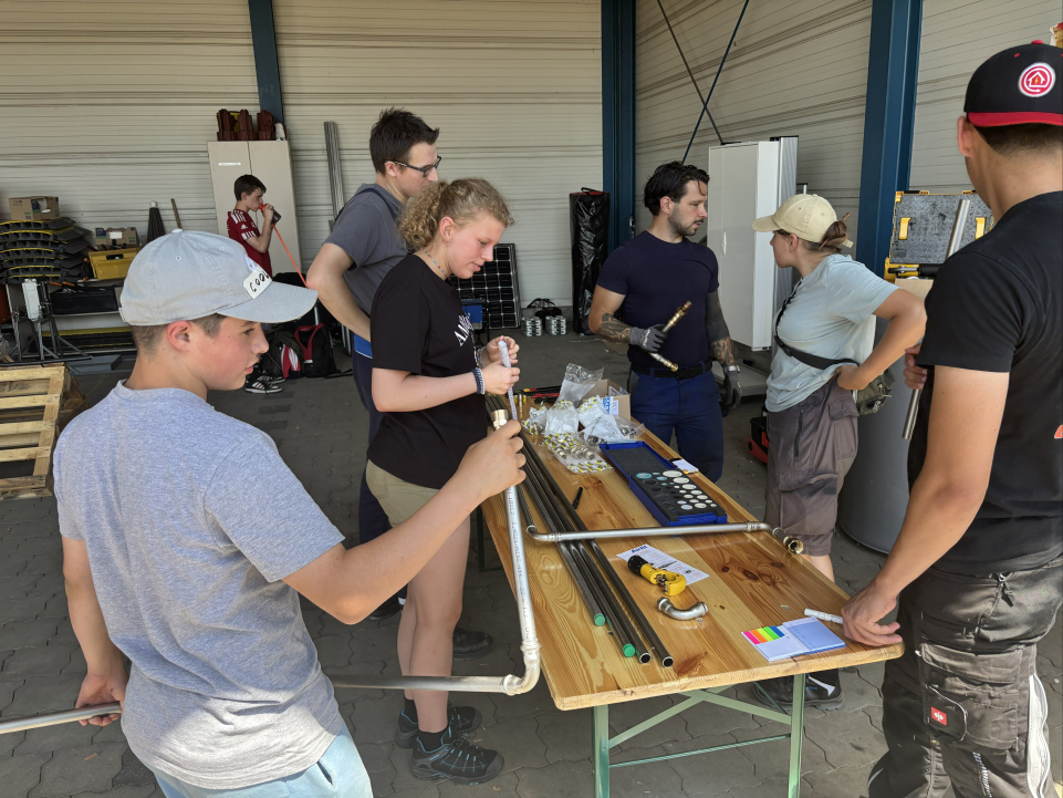 Eine Gruppe von Jugendlichen und Erwachsenen arbeitet in einer Werkstatt an einem Tisch mit Rohren, Verbindungsstücken und Werkzeugen. Im Hintergrund ist ein Solarmodul zu sehen.