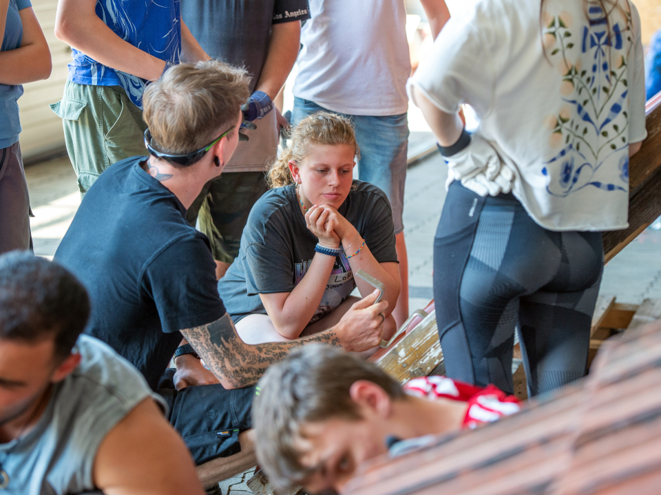 Mehrere junge Menschen sitzen und stehen in einem Innenraum an einem Schrägdach. Im Mittelpunkt unterhalten sich eine tätowierte Person mit dunklem Shirt und eine junge Frau, während weitere Personen um sie herumstehen.