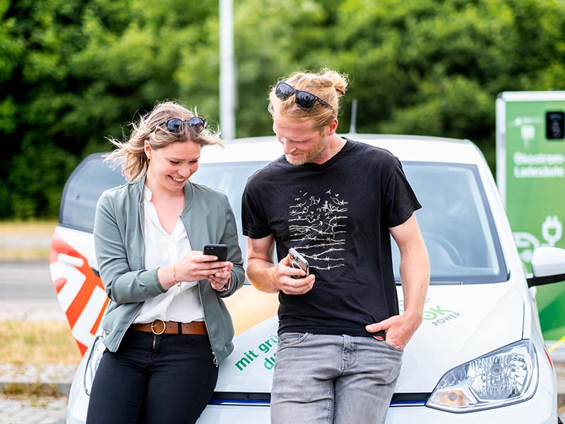 Junges Pärchen lehnt am Auto, schaut aufs Handy und lacht.
